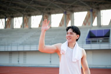Beyaz atletli Asyalı bir adam yarış pistinde dururken gülümsüyor ve elini sallıyor. Selamlama ya da hoşça kal, dışarıda egzersiz ya da eğitim.
