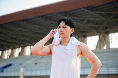 Beyaz atletli Asyalı adam ayakta ve terini havluyla siliyor, koştuktan sonra dinleniyor. Açık hava egzersizi ya da eğitim. Spor ve sağlık.