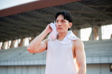 Beyaz atletli Asyalı adam ayakta ve terini havluyla siliyor, koştuktan sonra dinleniyor. Açık hava egzersizi ya da eğitim. Spor ve sağlık.