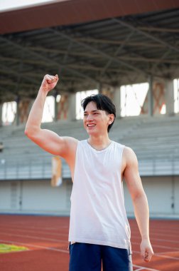 Beyaz kolsuz tişörtlü Asyalı adam yarış pistinde dururken gülümsüyor ve yumruğunu kaldırıyor. Bir koşuyu bitirmek ya da bir arkadaşa tezahürat etmek, açık hava egzersizi ya da eğitim.