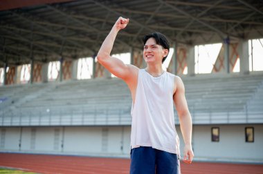 Beyaz kolsuz tişörtlü Asyalı adam yarış pistinde dururken gülümsüyor ve yumruğunu kaldırıyor. Bir koşuyu bitirmek ya da bir arkadaşa tezahürat etmek, açık hava egzersizi ya da eğitim.