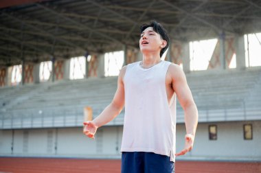 Beyaz tişörtlü Asyalı adam kolları açık ve derin bir nefes alıyor, koştuktan sonra dinleniyor. Açık hava egzersizi ya da eğitim. Spor ve sağlık.