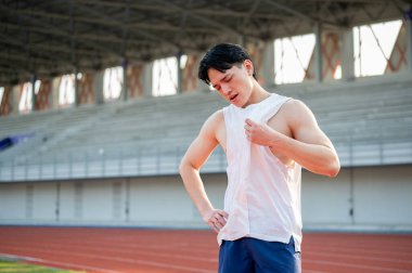 Asyalı bir adam bir eli belinde, diğer eli de beyaz atletle kendini yelpazeliyor. Koştuktan sonra sıcak ya da yorgun. Açık hava egzersizi ya da eğitim.
