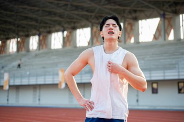 Asyalı adam bir eli belinde, diğer eli beyaz bir bluzu tutuyor, koştuktan sonra sıcak ya da yorgun hissediyor. Açık hava egzersizi ya da eğitim.