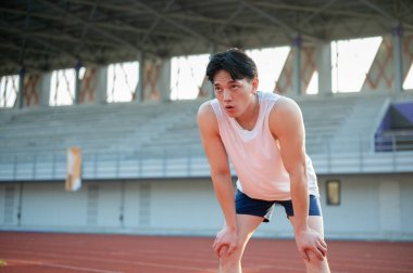 Beyaz kolsuz tişörtlü Asyalı adam elleri dizinde yarış pistinde koştuktan sonra dinleniyor. Açık hava egzersizi ya da eğitim. Spor ve sağlık.
