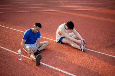 İki Asyalı atlet yarış pistinde bacak kaslarını esnetiyorlar. Koşmak ya da koşmak, dışarıda egzersiz yapmak ya da egzersiz yapmak, spor ve sağlık.