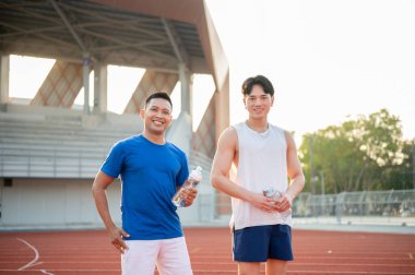 İki gülümseyen Asyalı sporcu su şişelerini tutuyor ve yarış pistinde birlikte duruyorlar. Koşmak, koşmak, dışarıda egzersiz yapmak ya da spor yapmak..