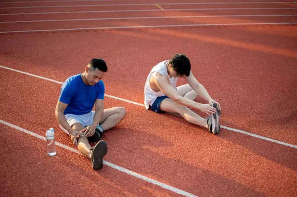 İki Asyalı atlet yarış pistinde bacak kaslarını esnetiyorlar. Koşmak ya da koşmak, dışarıda egzersiz yapmak ya da egzersiz yapmak, spor ve sağlık.