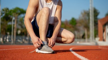 Spor ayakkabı bağcığını hipodromda bağlarken beyaz kolsuz tişörtlü bir adamın önünde çömelmek. Açık hava egzersizi, atlet eğitimi, spor ve sağlık..