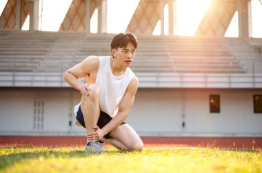 Beyaz atletli Asyalı adam çim tarlasında diz çökmüş, elleri dizinde ve ayak bileğinde. Acı ya da yaralanma. Açık hava egzersizi ya da eğitim. Spor ve sağlık.