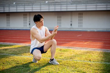 Beyaz kolsuz tişörtlü Asyalı adam çimlerin üzerinde çömeliyor ve bileğini tutuyor. Bilek ağrısı ya da yaralanma. Açık hava egzersizi ya da eğitim. Spor ve sağlık.