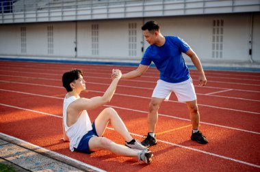 Asyalı adam arkadaşının elini tutuyor ya da çekiyor yarış pistinde düştükten sonra ayağa kalkmasına yardım ediyor. Açık hava egzersizi ya da eğitim. Spor ve sağlık.