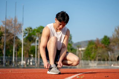 Beyaz kolsuz tişörtlü Asyalı adam spor ayakkabı bağcığını hipodromda bağlarken çömeliyor. Spor ya da spor, spor, spor ve sağlık..