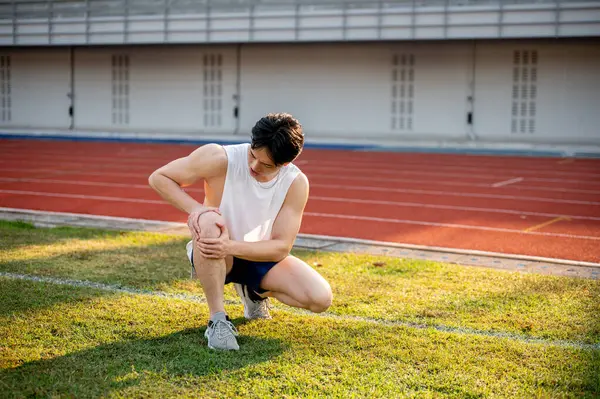 Beyaz kolsuz tişörtlü Asyalı adam çömelmiş ve dizini çimlerin üzerinde tutuyor. Diz ağrısı ya da yaralanma. Açık hava egzersizi ya da eğitim. Spor ve sağlık.