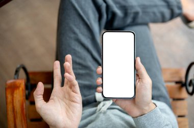 Bir elinde beyaz ekran akıllı telefonu, diğer elinde açıklayıcı bir jestle sandalyede bacak bacak üstüne atarak oturuyor. Boş ekran modeli, Ürün görüntüsü, Reklam.