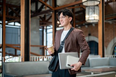 Kahverengi takım elbiseli Asyalı adam kafede elinde dizüstü bilgisayarla kahve fincanı tutarken ya ayakta duruyor ya da yürüyor. Çalışmak ya da dışarıda çalışmak.
