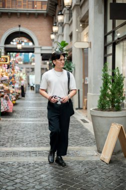 Beyaz tişörtlü Asyalı adam elinde kamerasıyla alışveriş merkezinin koridorunda ya da kapalı markette dolaşıyor. Fotoğraf gezisi. Turistik bölge.