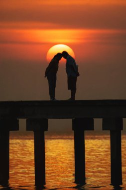Güneşin doğuşu ve deniz dalgalarının yansıması ile rıhtım köprüsünde romantik anlar yaşayan çiftlerin silueti.