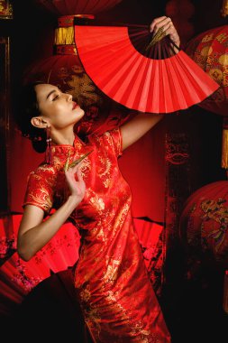 studio portrait of asian woman in tradional chinese red costume holding chinese fan with background of chinese lantern for chinese new year celebration, translation: wish you prosperity and wealth