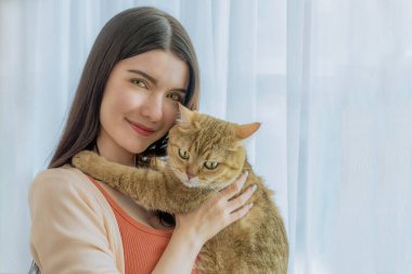 Genç bir Asyalı kadının sevgili kedisini sımsıkı tutması çok iç açıcı bir sahne. Parlak bir pencerenin yanında duruyorlar, sabah ışığı içeri giriyor ve evdeki bu samimi ana sıcaklık katıyor.