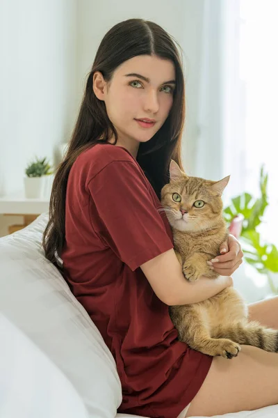 The portrait showcases a beautiful young Asian woman embracing her cute cat tightly as they share a heartwarming moment in the morning, sitting on the bed at home