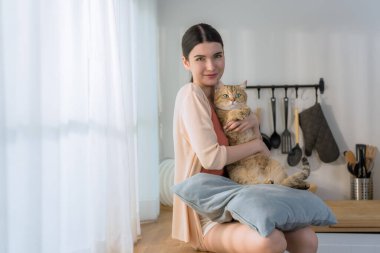 Çekici genç bir Asyalı kadın mutfak tezgahında oturuyor, sabah güneşi gibi sevimli kedisini sıkıca tutuyor.