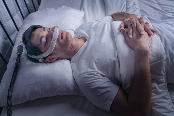 Happy and healthy Asian man wearing Cpap mask sleeping smoothly in bed all night without snoring. Obstructive sleep apnea therapy