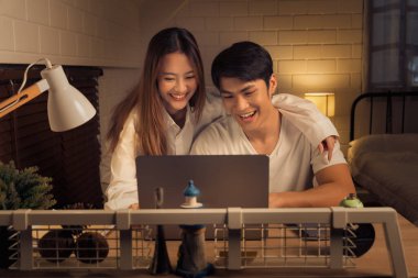 Genç çift sıcak ışıklandırmalı sıcak bir odada dizüstü bilgisayar kullanırken keyifli bir anı paylaşıyor.