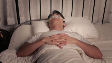 Barışçıl Asyalı erkek uyku apnesi ve horlama tedavisi için CPAP maskesi ve makine kullanarak uyuyor.