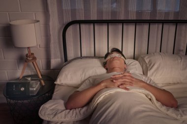 Huzurlu Asyalı erkek CPAP maskesiyle uyuyor, cihaz komodinin üzerinde görünür, uyku apnesi ve horlama tedavisi için.
