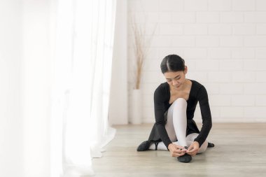 Siyah elbiseli bir balerin ahşap bir zeminde oturur. Dikkatlice dans ayakkabılarını parlak, havadar bir zemine yaslar.