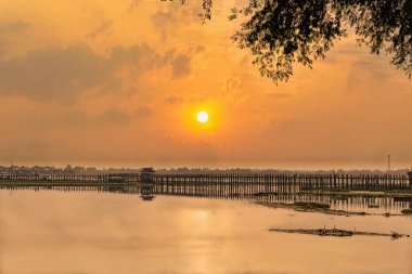 Altın gün doğumu Mandalay, Myanmar 'daki U-Bein Köprüsü' nün üzerinde sakin bir ışık saçıyor ve aşağıdaki sakin sularda yansımalar parlıyor.