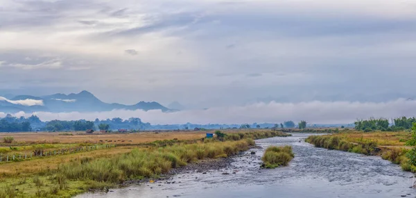 The serene waters of a river in Putao flow gently amidst fields, with the ethereal beauty of mist-shrouded mountains looming in the distance under a soft morning sky
