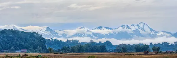 The panoramic vista of Putao unfolds a tranquil rural scene against the backdrop of Myanmar\'s majestic, snow-covered mountains, veiled in misty clouds
