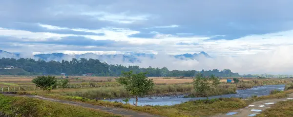 The serene waters of river in Putao flow gently amidst fields, with ethereal beauty of mist-shrouded mountains looming in distance under soft morning sky