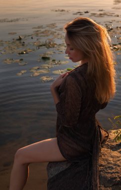 Güzel romantik bir kadın göl kenarında oturuyor. Gün batımında güneşin altında iç çamaşırlarıyla ıslak bir kadın. Doğal güzellik, uzun saç.