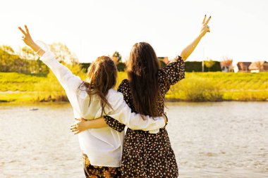 Yazın gün batımında birlikte bir yolculuk sırasında nehre bakan elleri gökyüzüne bağlı iki genç kadın.