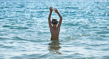 Siyah şnorkelli beyaz bir çocuk beline kadar suya batmış ve ellerini havada tutuyor. Bir çocuk denizde yüzer. Açık hava eğlencesi.