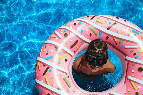 Water entertainment with a pink inflatable circle in the pool. A boy in a swimming mask looks up. View from above.