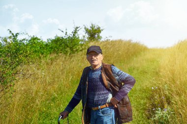 Bıyıklı ve gözlüklü gülümseyen yaşlı adam çimenli bir yolda duruyor, baston tutuyor ve kolsuz koyun derisi ceket giyiyor. Dedemin portresi güneşli bir zemine kurulmuş ve kameraya bakıyor..
