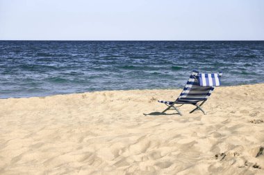 Kumlu bir sahilde, mavi ve beyaz çizgili plaj sandalyesi. Huzurlu bir sahilde yalnızlık.
