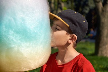 Güneşli bir bahar gününde pamuk helva bulutunun tadını çıkaran şapkalı bir çocuk. Çocuğun neşeli ifadesi ve pamuk helvanın canlı renkleri çocukluk mutluluğu hissi yaratıyor..