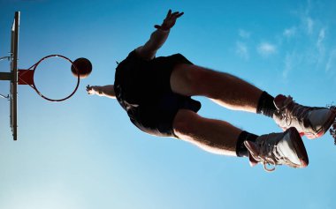 Genç adam ringde bir gol atıyor, arka planda açık mavi gökyüzü olan bir sahada şut atıyor. Sokak basketbolu.