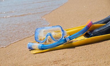 Bir çift parlak sarı palet ve mavi şnorkel tropik bir plajın yumuşak kumlarında uzanıyor. Turkuaz okyanus dalgaları arka planda yavaşça kıyıya vurur..
