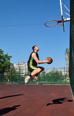 Basketbol oynayan genç bir adam arka planda açık mavi gökyüzü olan bir sahada şut atıyor. Sokak oyunu.