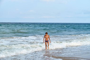 Sahildeki dalgaların tadını çıkaran genç bir çocuk, engin mavi okyanusla sahil boyunca yürüyordu açık bir gökyüzünün altında ufka doğru uzanan.