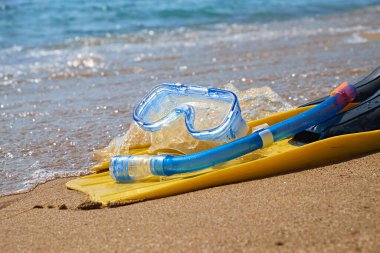 Bir çift parlak sarı palet ve mavi şnorkel tropik bir plajın yumuşak kumlarında uzanıyor. Turkuaz okyanus dalgaları arka planda yavaşça kıyıya vurur..