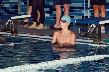 Yüzme şapkası ve gözlüklü genç bir yüzücü kapalı bir havuzun kenarında yüzmeye hazırlanıyor. Odaklanmış görünüyor, muhtemelen bir yüzme yarışması ya da antrenman sırasında..