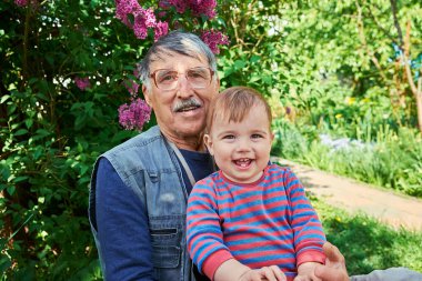 Gözlüklü ve bıyıklı yaşlı bir adam kucağında mutlu, gülümseyen bir bebek tutuyor. Canlı yeşil yaprakları ve çiçek açan mor çiçekleri olan bir bahçede oturuyorlar..