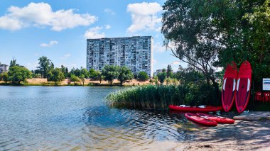 Sahilde kırmızı kanoları ve kürekleri olan, yemyeşil ve açık mavi gökyüzüyle çevrili, sakin bir göl kenarındaki modern apartman binası, huzurlu bir deniz manzarası yaratıyor..